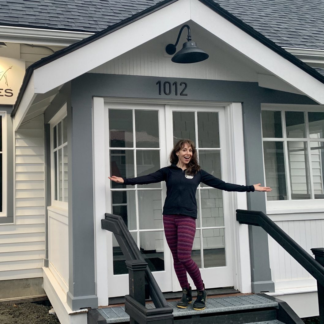 A woman standing on the front steps of a small house near a Juneau Pilates Studio.